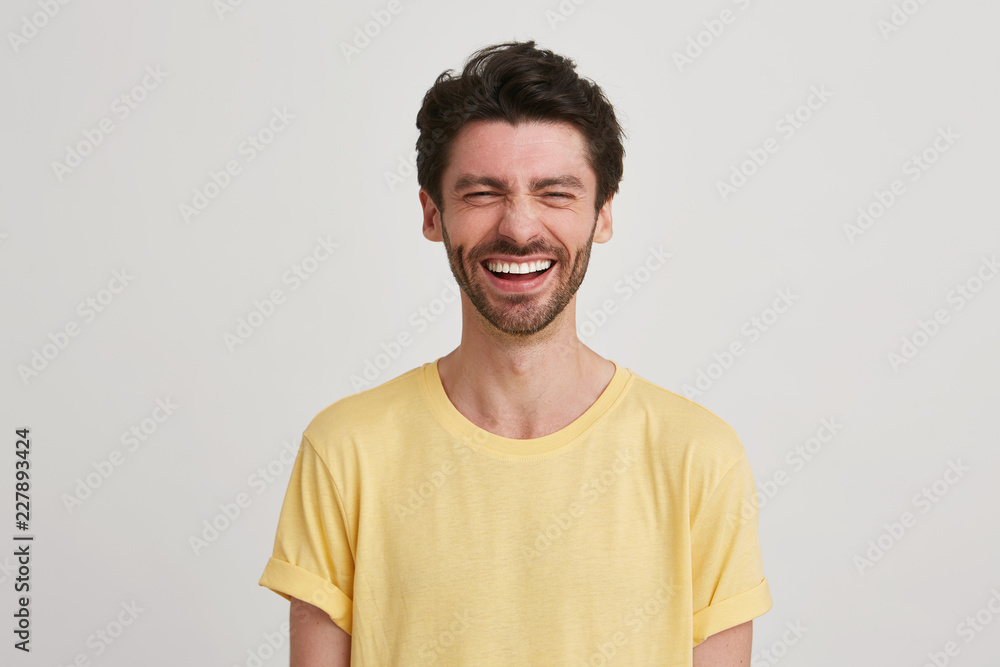 Close up of laughing happy screwing up his eyes attractive young man with dark brown hair and beard wears yellow tshirt, isolated over white background Close up of laughing happy screwing up his eyes attractive young man with dark brown hair and beard wears yellow tshirt, isolated over white background