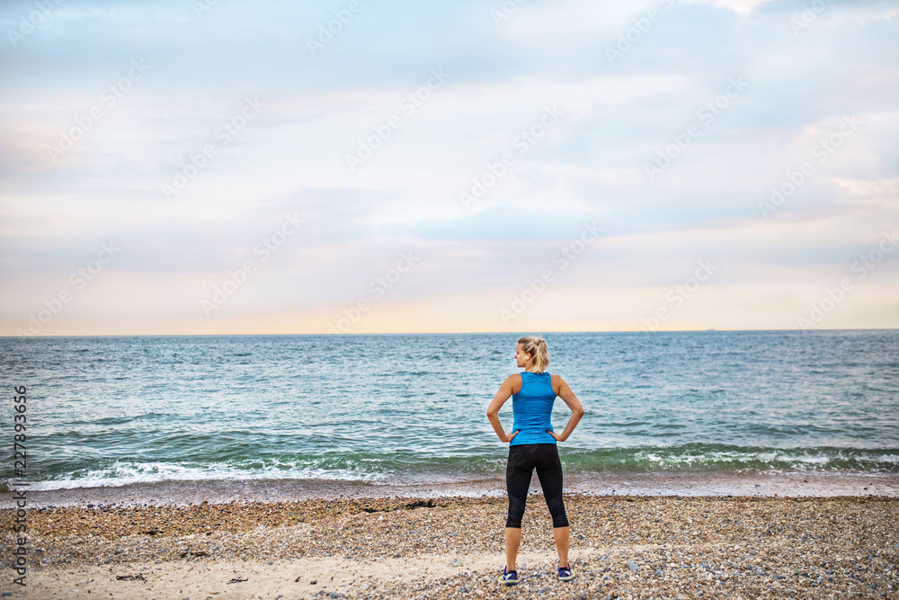 Rear view of young sporty woman runner in blue sportswear standing on the beach.