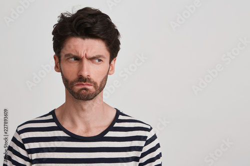 Young man with dark brown hair and beard wears black and white striped tshirt, looks angry, lips pursed frowning brows, head tuned rightside, stands leftside isolated over white background photo