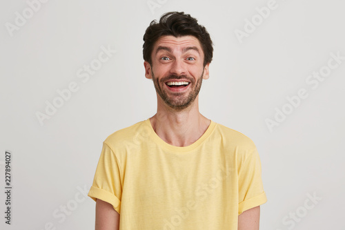 Close up of happy and very excited attractive young brunet with beard eyebrows raised wears yellow casual tshirt, isolated over white background