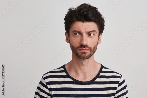 Young man with dark brown hair and beard wears black and white striped casual tshirt looks thinking, a little frowning brows, sight turned leftside, isolated over white background photo