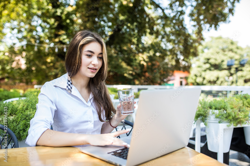 Pretty girl drinking water in restaurant. Beautiful girl in white shirt working on her laptop in coffee shop. Close up lady hand typing on computer at cafe Pretty girl drinking water in restaurant. Beautiful girl in white shirt working on her laptop in coffee shop. Close up lady hand typing on computer at cafe