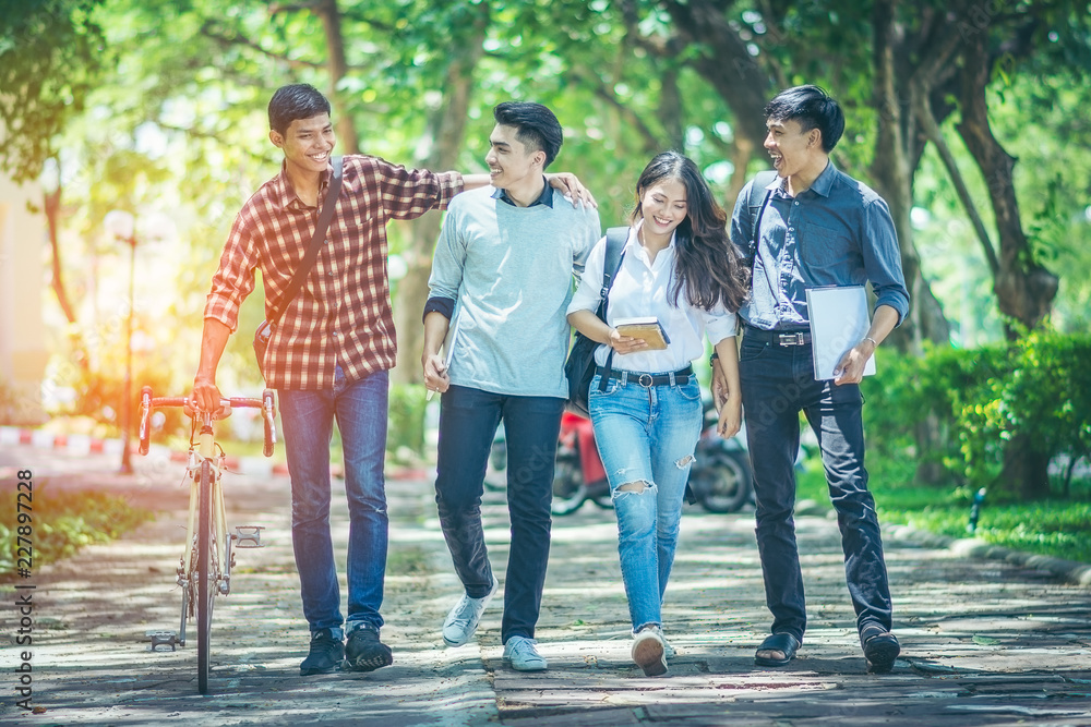 Fototapeta premium A group of Asian student was walking on the road within the University. They talk to smile happily and motivate the bicycle to walk together. In the shade of trees streets and the sun in the afternoon