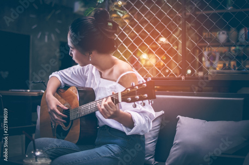 Asian girls wearing white shirts and jeans. She is sitting on a black couch is currently vacationing by playing guitar in a coffee shop. Iced coffee is placed on a wooden table with copy space.