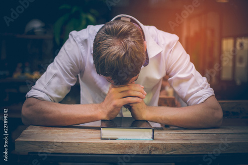 The young man handsome caucasian Americans handsome man sitting with hands clasped in prayer for blessings from God. A Bible rested on a wooden table with copy space.