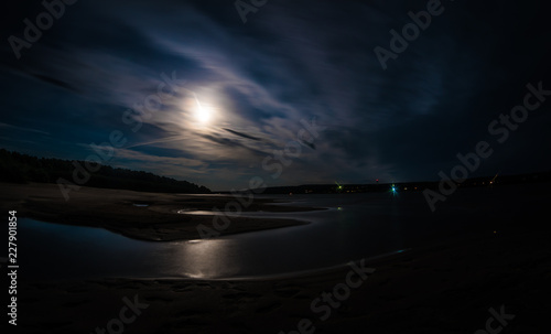 Obraz na plátně A long exposure shot of Tom river with moonlit water and shores under the cloudy night sky, foot-marks on sand