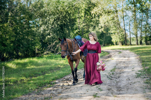 A woman with a bouquet of flowers dressed in a long burgundy dress with sleeves leads a brown horse along a rural road
