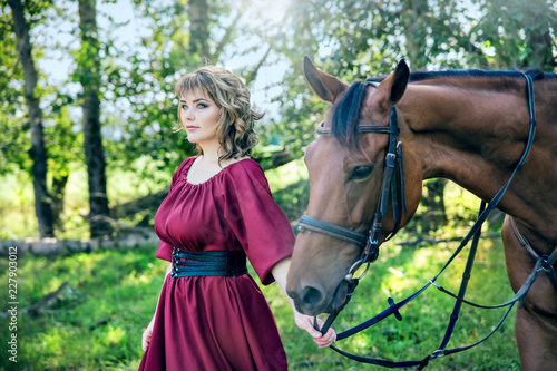 A woman with a bouquet of flowers dressed in a long burgundy dress with sleeves leads a brown horse on the green grass on the background of trees.