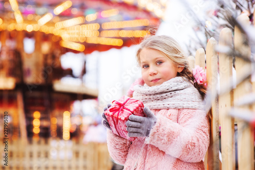 happy child girl holding christmas gift outdoor on the walk in snowy winter city decorated for new year holidays.