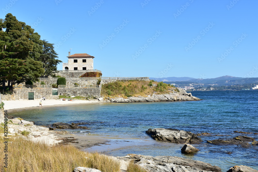 Small beach in a bay with promenade, rocks, trees and a house on a hill. Turquoise and clear water, blue sky, sunny day. Rias Baixas, Galicia, Spain. 