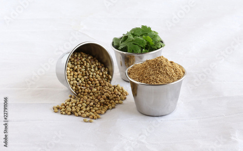 Coriander powder, plucked coriander leaves, and spilled coriander seeds, in steel bowls on a crumpled paper background.