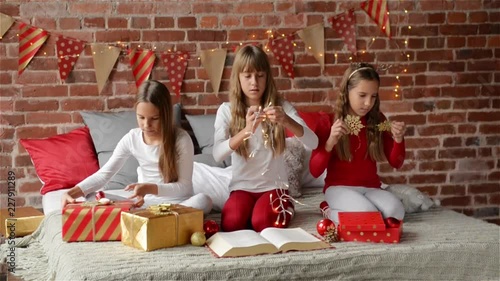 Three sisters dressed in xmas pajamas sitting on a decorated bed for the Christmas, triplet girls opening presents, Merry Christmas and Happy New Year