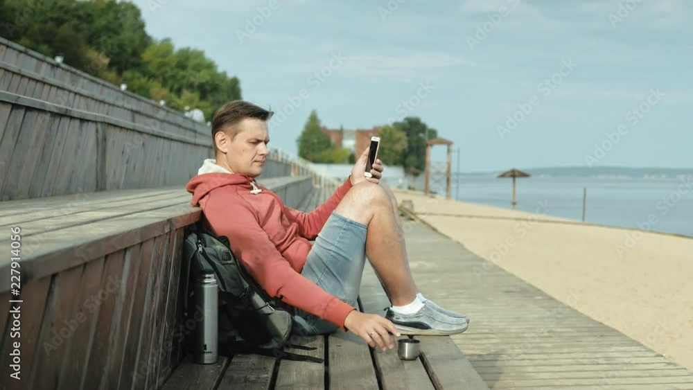 Mature man, tourist using a smartphone, sitting on the beach on a wooden bench Mature man, tourist using a smartphone, sitting on the beach on a wooden bench
