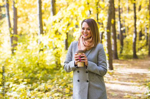 Autumn, nature, people concept - young woman in a blue coat standing in the park on a background of trees with a cup of coffee. © satura_