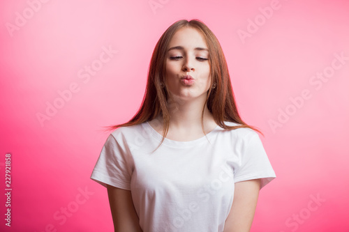Cute lovely young woman in white shirt standing and sending kiss over pink background. Girl looking down