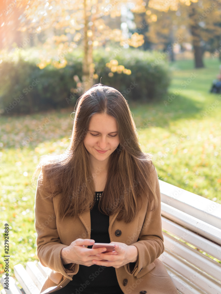 Beautiful teen girl sitting in the Park on a bench, holding a smartphone and chatting online on the Internet. Woman with long hair Beautiful teen girl sitting in the Park on a bench, holding a smartphone and chatting online on the Internet. Woman with long hair