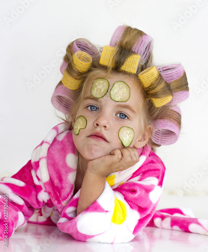 Девочка в банном розовом халате с бигудями и кусочками огурцов на лице.