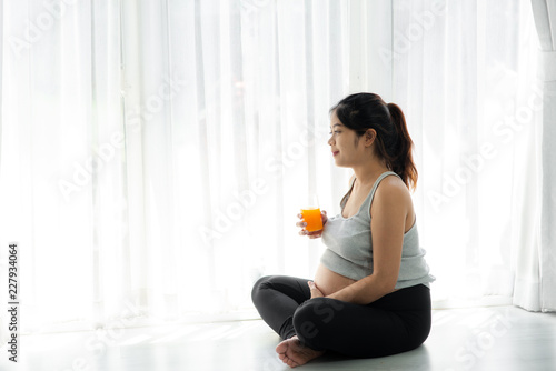Asian pregnant woman holding a glass of orange juice and sitting near window, lifestyle concept.