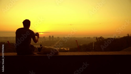 A man photographs the beautiful landscape that he watchs with his dog. The man wears a striped T-shirt, it's the sunrise.