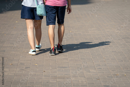 On a sunny day, a couple of people walk along the sidewalk. The human shadows are visible on the sidewalk. View from the back.