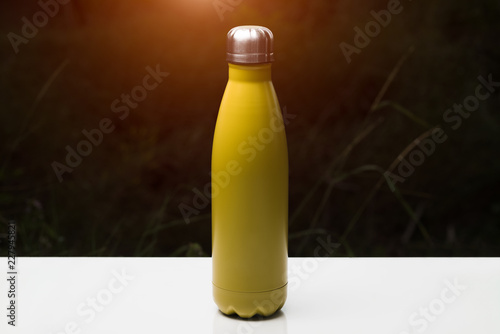 Stainless thermo bottle for water, tea and coffe, on white table. Dark grass background with sunlight effect. Thermos yellow color.