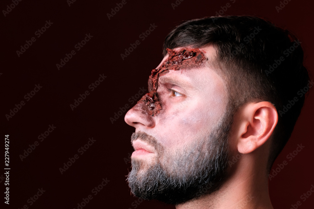 Obraz premium Profile of a young man with ragged facial wounds. A guy with Halloween make-up looks to the left against a burgundy background. Close-up. Copyspace.