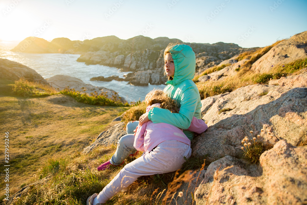 Two little girls play on rocky northern seashore. Sit, laugh, hug, explore the coastal rocks. Travel and enjoy a great adventure in Norway. Beautiful view of fjord and mountains in sunset. Two little girls play on rocky northern seashore. Sit, laugh, hug, explore the coastal rocks. Travel and enjoy a great adventure in Norway. Beautiful view of fjord and mountains in sunset.