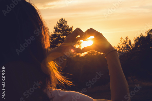 Woman makes heart with hands in sunset. 