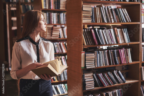 Young attractive student librarian reading a book between library bookshelves photo