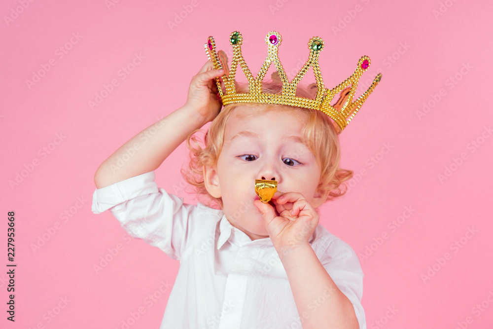 little blond boy with golden crown head curls hairstyle 4-5 year old in studio on pink background blowing noisemakers horn-whistle a birthday party celebrates Christmas and New Year king of the party