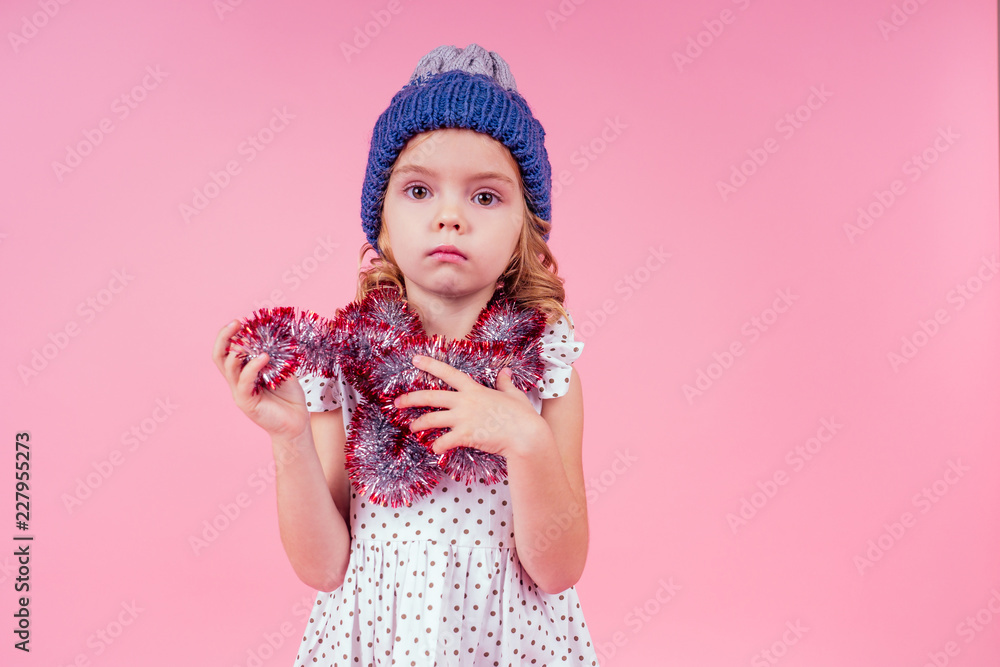 Beautiful little girl in dress and blue knitted hat in studio on white background. Happy little smiling female child with shiny Christmas tinsel. Christmas miracles Happy New Year Beautiful little girl in dress and blue knitted hat in studio on white background. Happy little smiling female child with shiny Christmas tinsel. Christmas miracles Happy New Year