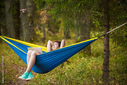 Image of young woman with hands behind head resting in hammock with laptop in forest