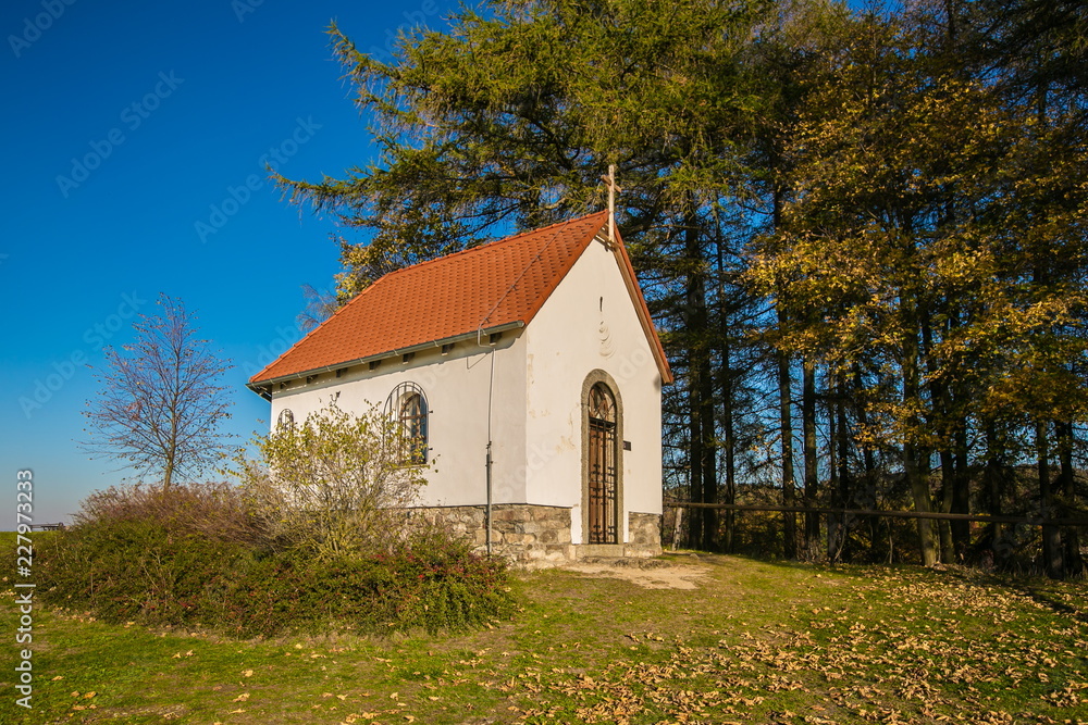 Autumn landscape, side view of white chapel of Saint Mary Magdalene with red roof, wooden cross on top of front wall, iron bars on door, bright sunny day, clear blue sky, trees, dry leaves on grass Autumn landscape, side view of white chapel of Saint Mary Magdalene with red roof, wooden cross on top of front wall, iron bars on door, bright sunny day, clear blue sky, trees, dry leaves on grass