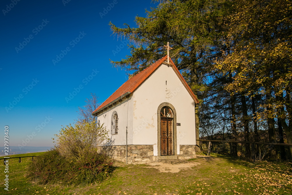 Autumn landscape, view of white chapel of Saint Mary Magdalene with red roof, wooden cross on top of front wall, iron bars on door, bright sunny day, clear blue sky, trees, grass, dry leaves on ground Autumn landscape, view of white chapel of Saint Mary Magdalene with red roof, wooden cross on top of front wall, iron bars on door, bright sunny day, clear blue sky, trees, grass, dry leaves on ground