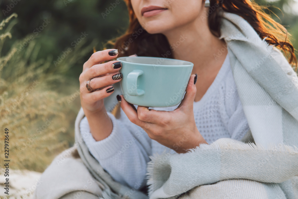 Young woman is sitting on the beach in warm plaid and drinking coffee from big cup. Young woman is sitting on the beach in warm plaid and drinking coffee from big cup.