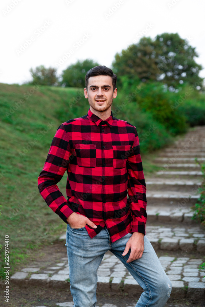Portrait of stylish male student with bread posing outdoors, looking at camera. Handsome man wearing in red checked shirt walking in autumn park. Concept of study, and mans fashion. Portrait of stylish male student with bread posing outdoors, looking at camera. Handsome man wearing in red checked shirt walking in autumn park. Concept of study, and mans fashion.