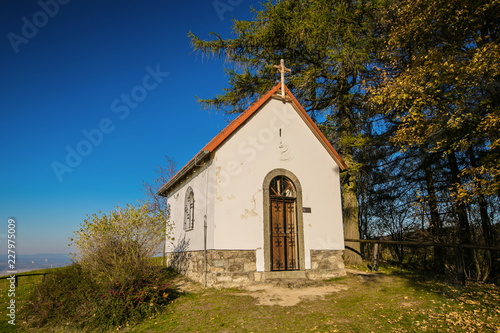 Autumn landscape, view of white chapel of Saint Mary Magdalene with red roof, wooden cross on top of front wall, iron bars on door, bright sunny day, clear blue sky, trees, grass, dry leaves on ground