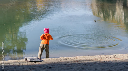 Photos A little girl, a child, in warm clothes and a red cap throws stones into the wat
