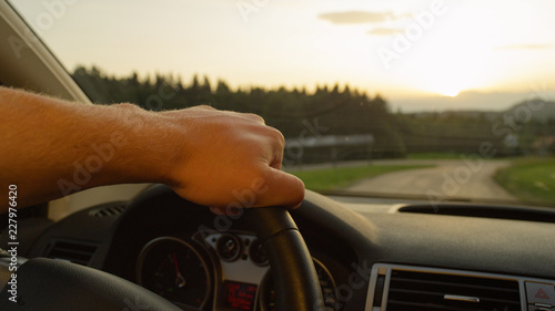 Fototapeta Naklejka Na Ścianę i Meble -  CLOSE UP: Unrecognizable man holds the steering wheel while driving at sunset