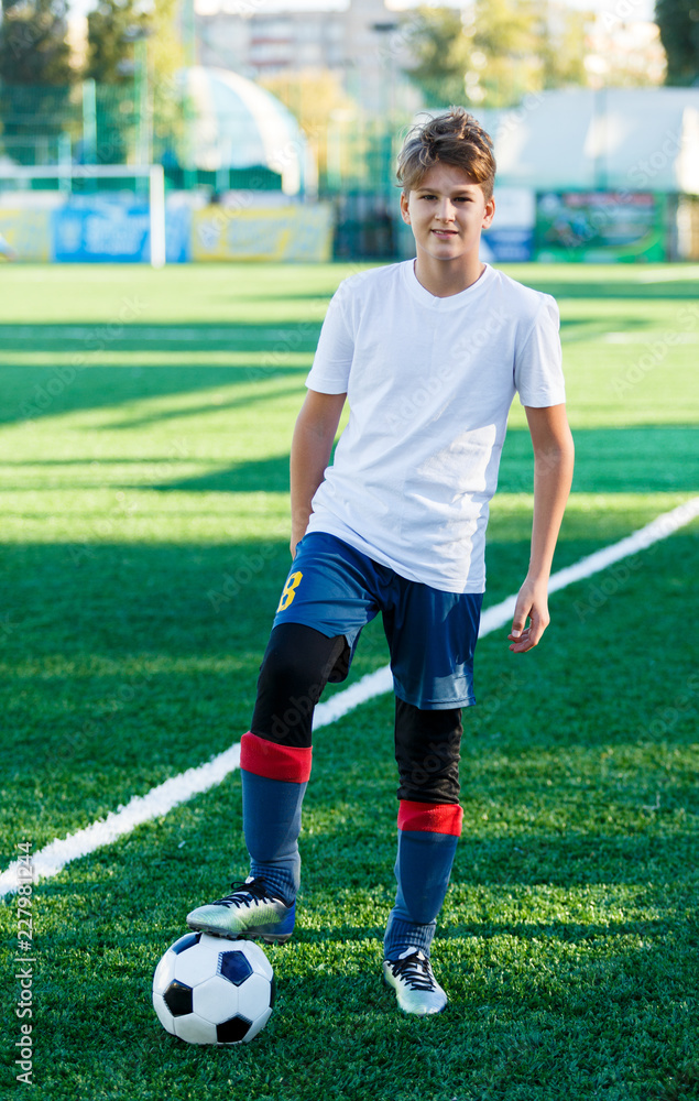 cute young boy in white blue sportswear stands next to classical black and white football ball on the stadium field. Soccer game, training, hobby concept. 