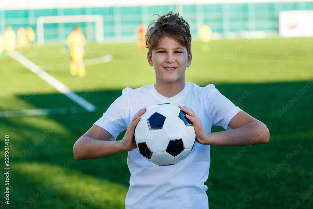 cute young boy in white blue sportswear holds classical black and white football ball on the stadium field. Soccer game, training, hobby concept. 