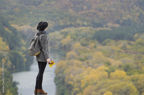 Girl with a backpack and a hat standing on a hill. Hands raised up. River and mountains below. Side view