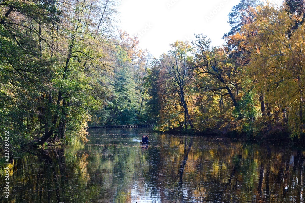 Fototapeta premium Ukraine. Kyiv. Pushcha Voditsa. Autumn landscape with trees reflecting in the water of the lake and people boating.