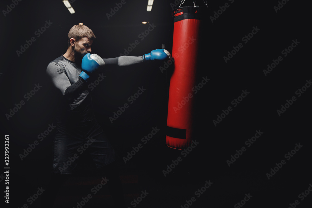 Young male boxer hitting punching bag on black background in gym.