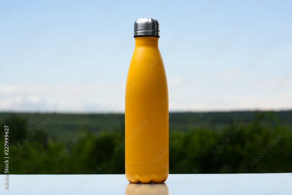 Thermo stainless bottle for water, tea or coffee. Sky and forest on background. On the glass desk. Thermos of matte yellow color. Thermo stainless bottle for water, tea or coffee. Sky and forest on background. On the glass desk. Thermos of matte yellow color.