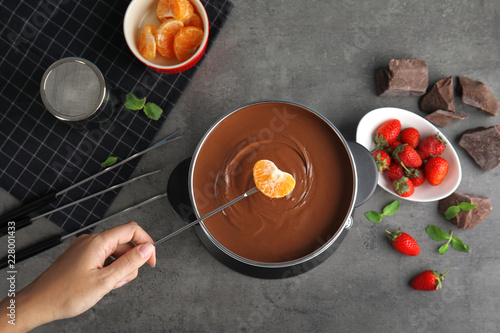 Woman dipping tangerine into pot with chocolate fondue on gray background, flat lay