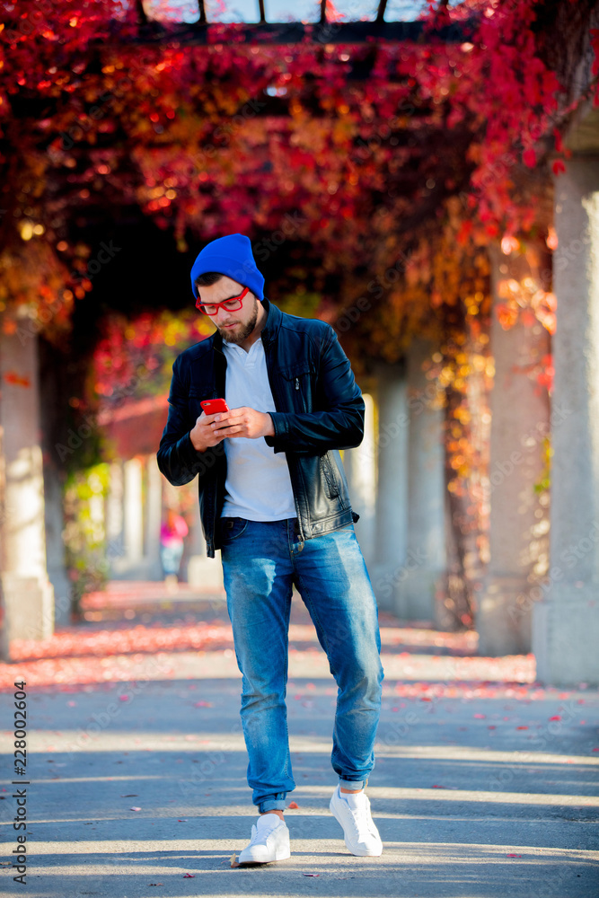 Young white guy using a mobile phone in a park with yellow trees on background. Autumn season time