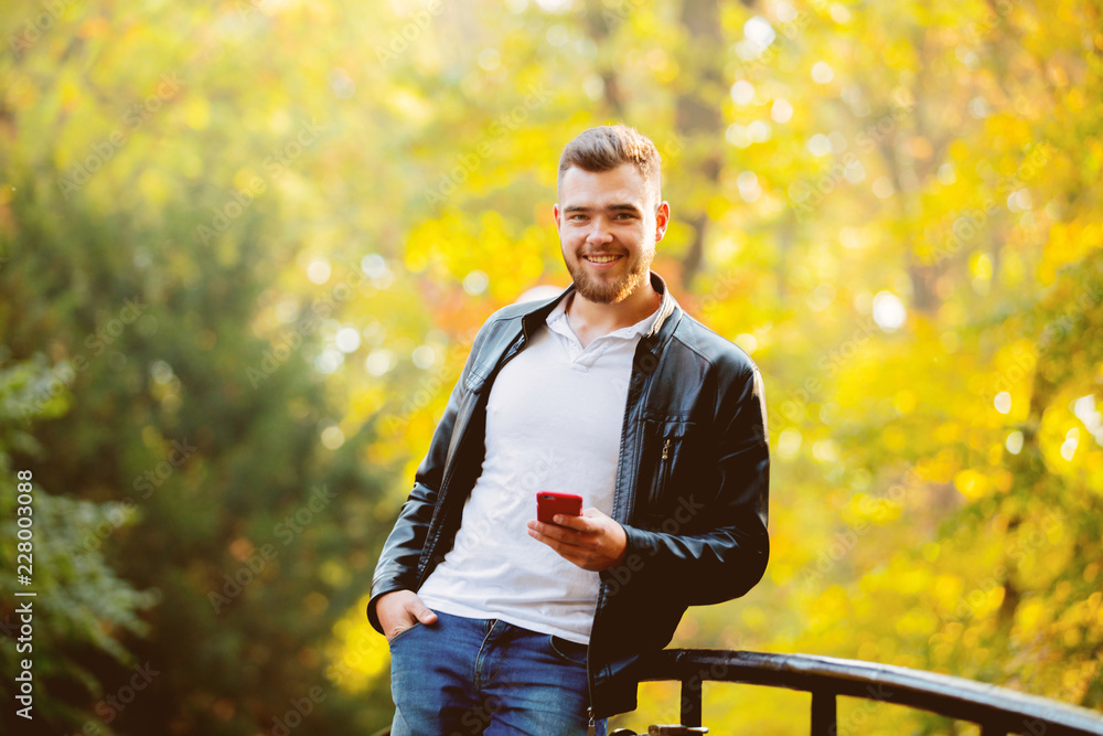 Obraz premium Young white guy using a mobile phone in a park with yellow trees on background. Autumn season time