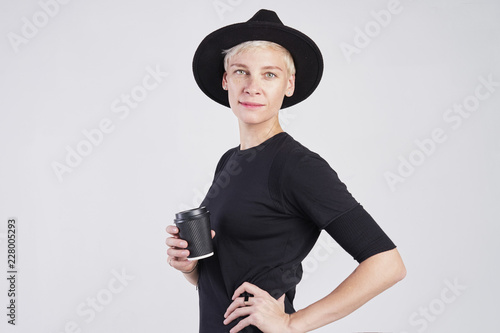 Portrait of blond caucasian woman wearing black clothes and hat having disposable paper cup of coffee, posing on white background. © olgapogorelova