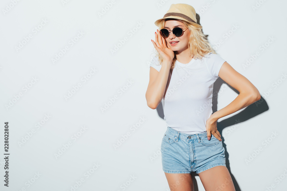 Portrait of a smiling attractive woman in sunglasses, summer clothes and hat posing while standing isolated over white background Portrait of a smiling attractive woman in sunglasses, summer clothes and hat posing while standing isolated over white background